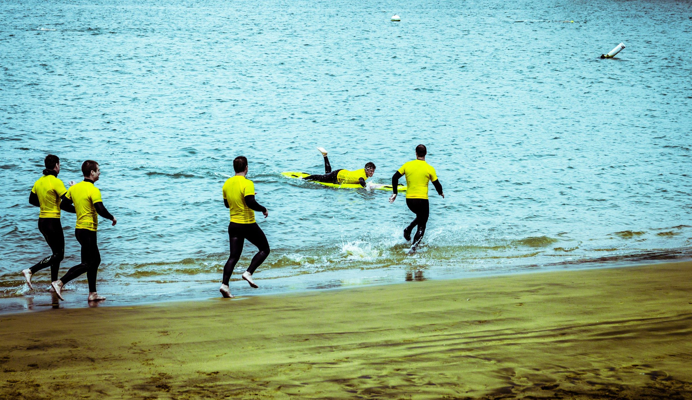 Des sauveteurs en lycra jaune entrent dans l’eau en courant, l’un d’eux déjà allongé sur sa planche