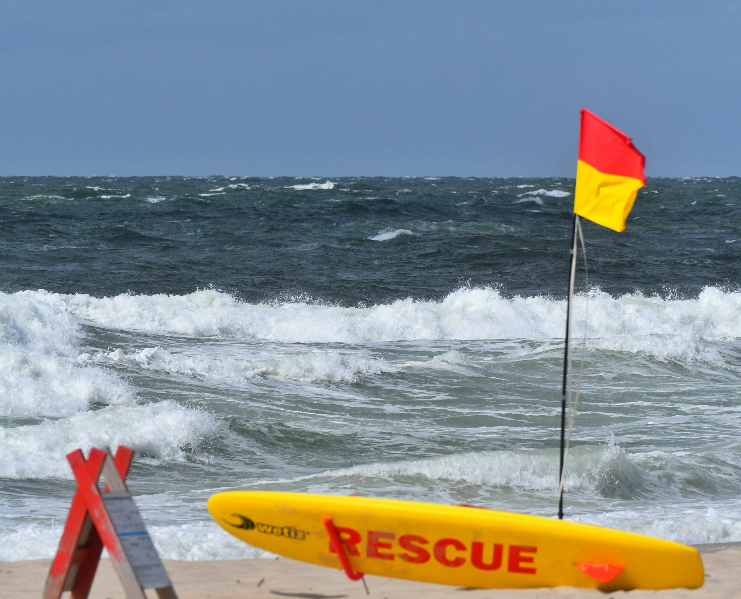 Une planche de sauvetage jaune « RESCUE » sur le sable, drapeau rouge et jaune devant une mer agitée